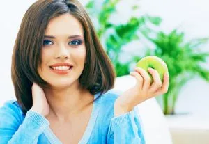 A woman with long dark hair smiling and holding a green apple, symbolizing dental health and strength.