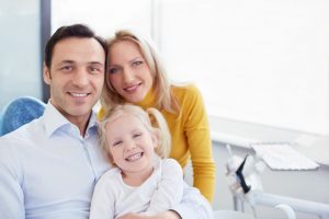 A happy family of three (mother, father, and young daughter) smiling together inside a bright dental office.