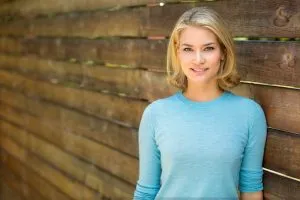 A portrait of a blonde woman in a light blue sweater smiling confidently while leaning against a rustic wooden wall.