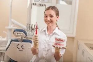 A female dental professional in a white coat smiling while holding a toothbrush and a dental model of human teeth.