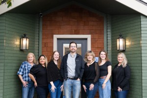 A professional group portrait of a dental team standing outside their office entrance. A male dentist is positioned in the center, flanked by six female staff members on either side. They are all smiling and dressed in casual professional attire, posed against a building with green brick and wood shingle siding.